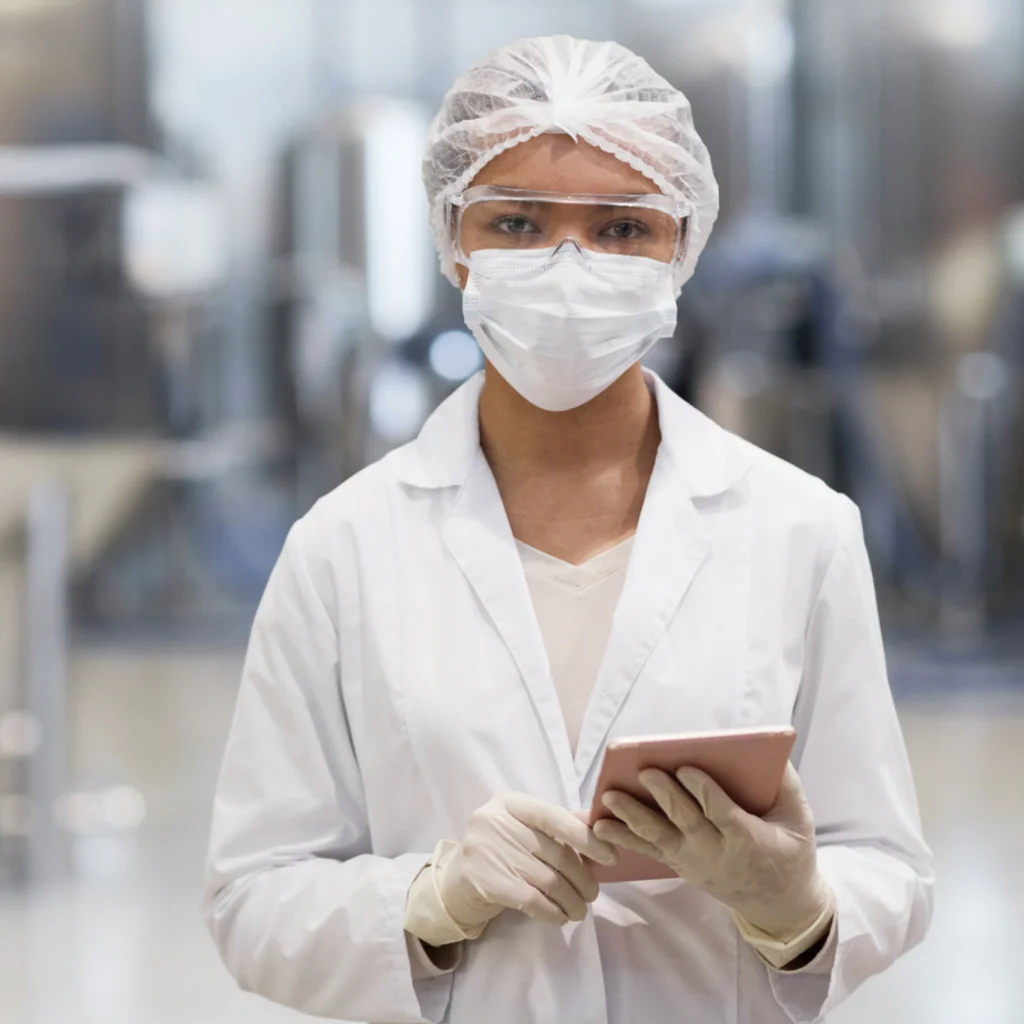 Eine Frau steht in einer Produktion mit einem Kittel, Atemschutzmaske und Klemmbrett