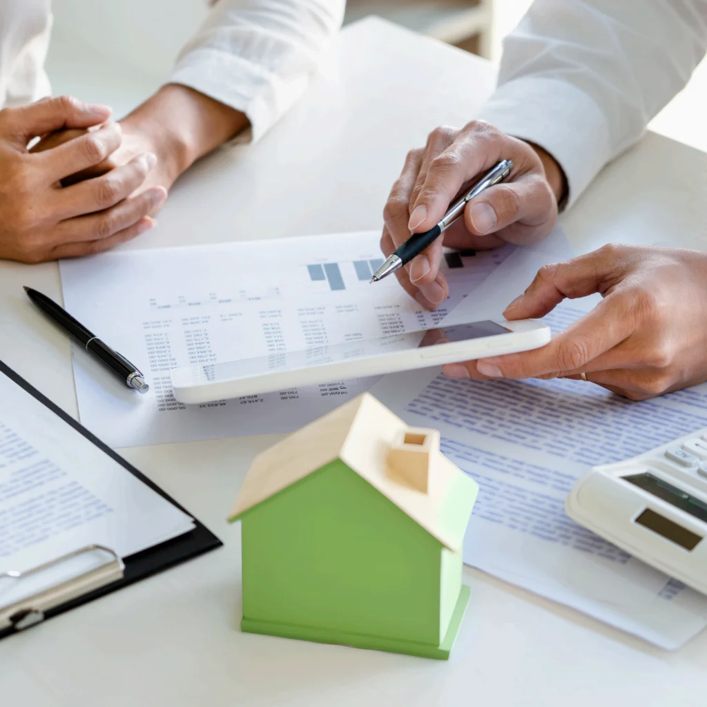 Real Estate Management auf Zeit Zwei Personen sitzen an einem Tisch mit Dokumenten und einem kleinen gruenen Holzhaus. Von den Personen sind nur die Unterarme jeweils mit weissen Hemd und Haende zu sehen.