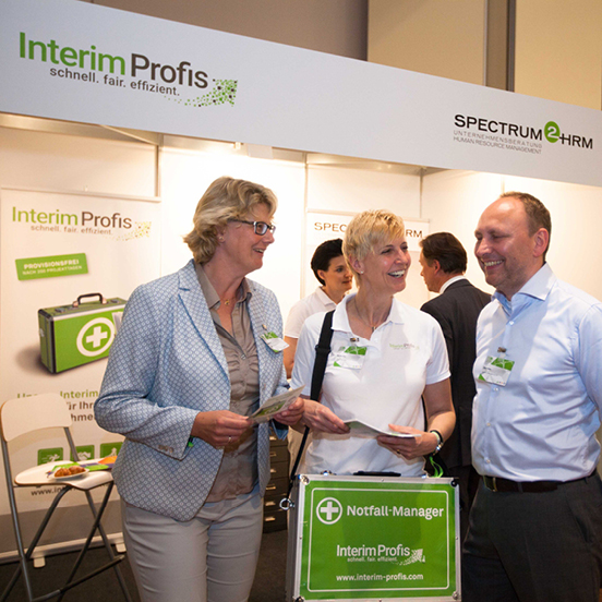 Netzwerk der Interim Profis - Partner werden Annette Elias und ein Interim Manager und eine Interim Managerin stehen an einem Messestand der Interim Profis