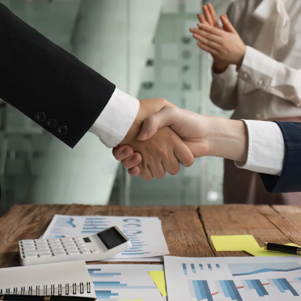 Arbeitsmarkt für Produktionsleiter Zwei Personen reichen sich ueber einen Tisch mit diversen Papieren die Haende. Dahiner steht eine weitere Person die sich in die Haende klatscht.