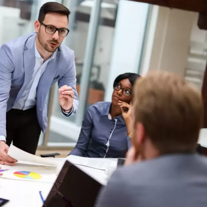 Interim Lean Manager Eine Gruppe von drei Personen sind in einem Buero, zwei Personen sitzen und Mann steht und spricht zu den beiden anderen Personen