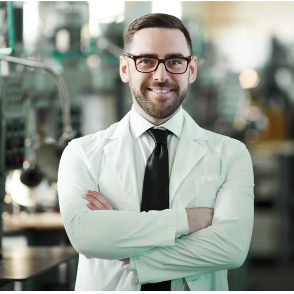 Ein Mann mit Brille, in weisser Laborkleidung und schwarzer Krawatte steht mit verschraenkten Armen in einer Produktionshalle und laechelt ins Bild