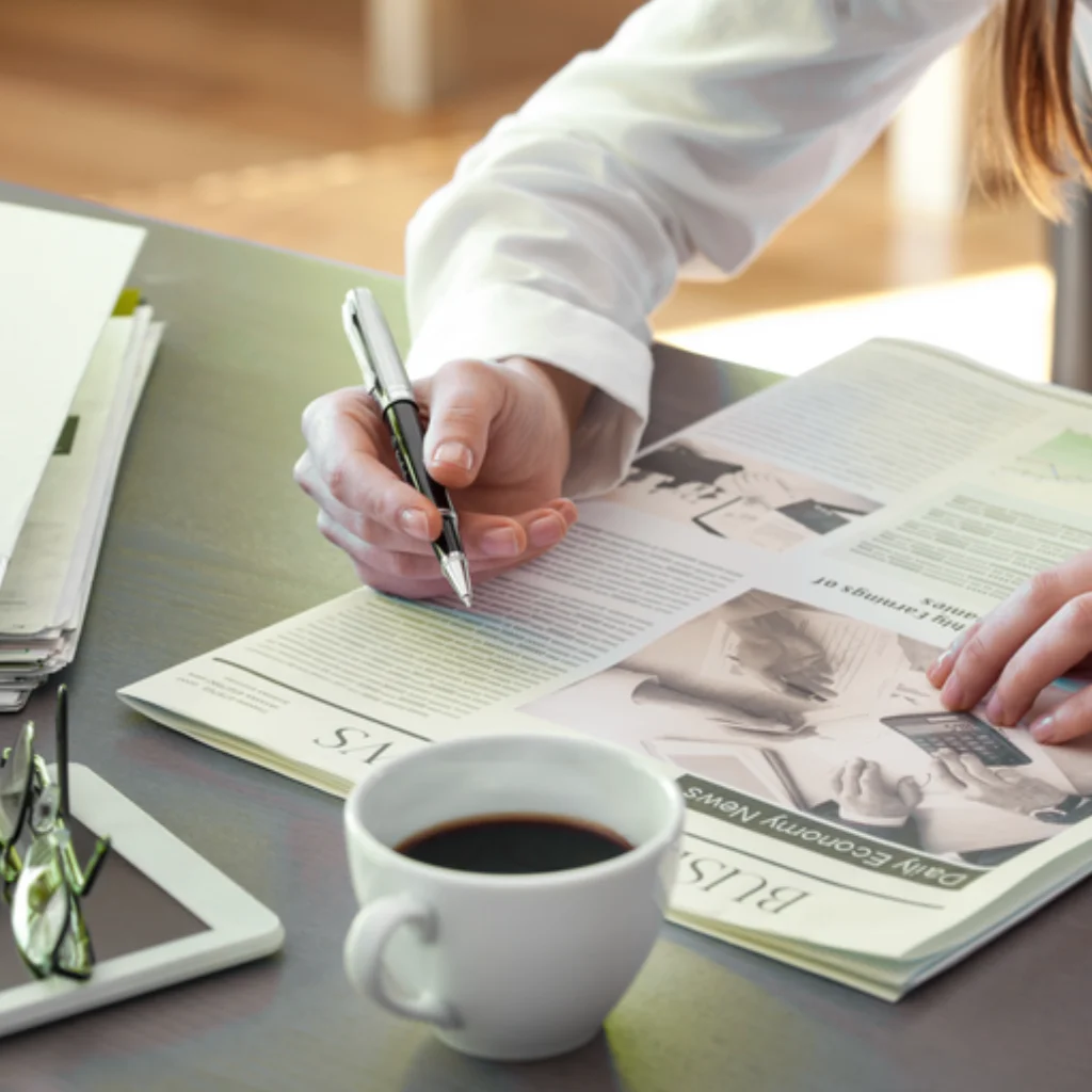 Eine Person sitzt an einem Schreibtisch und liest eine Zeitung, daneben steht eine Tasse Kaffe, eine Brille und Tablet