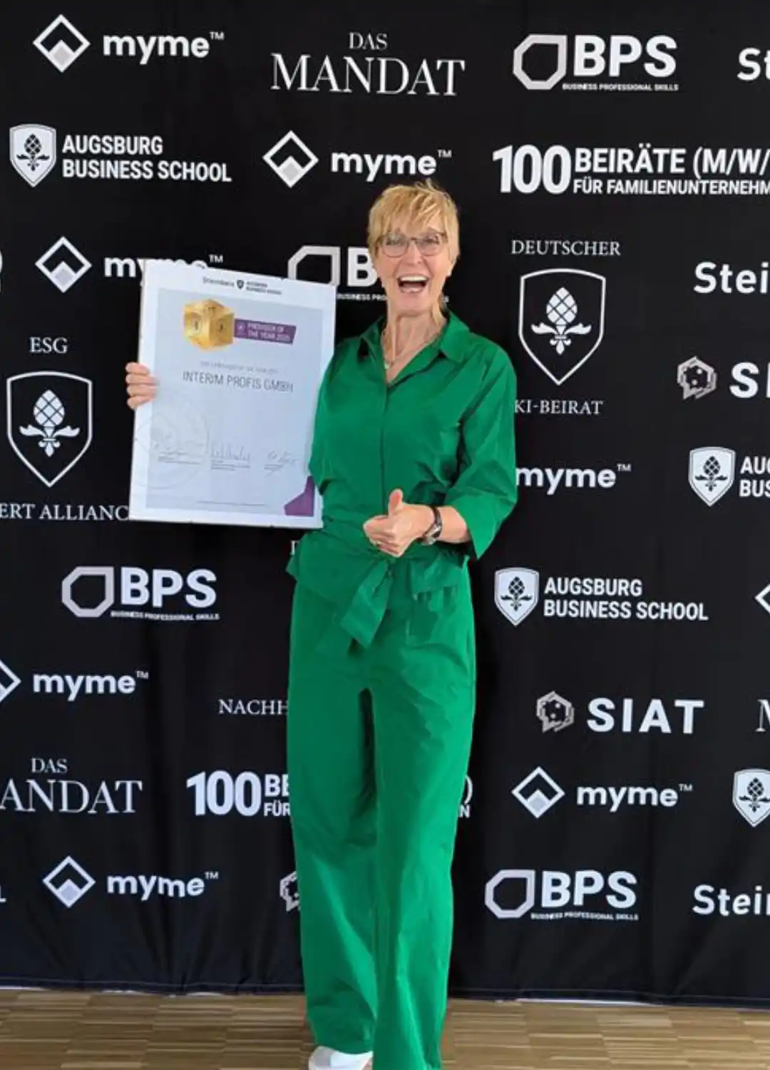 Annette Elias posiert mit dem Zertifikat ,,Interim Provider of the year 2025'' ausgestellt für die Interim Profis vor einer Fotowand mit den Sponsoren der Veranstaltung