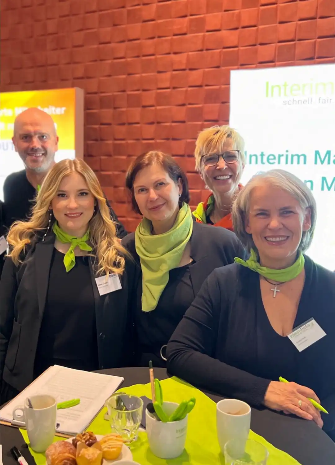Mitarbeitende der Interim Profis mit Magnus Barth, Eileen Dross, Kathrin Scholz.Arps, Annette Elias und Caroline Pusch stehen an einem Hochtisch mit Schreibutensilien