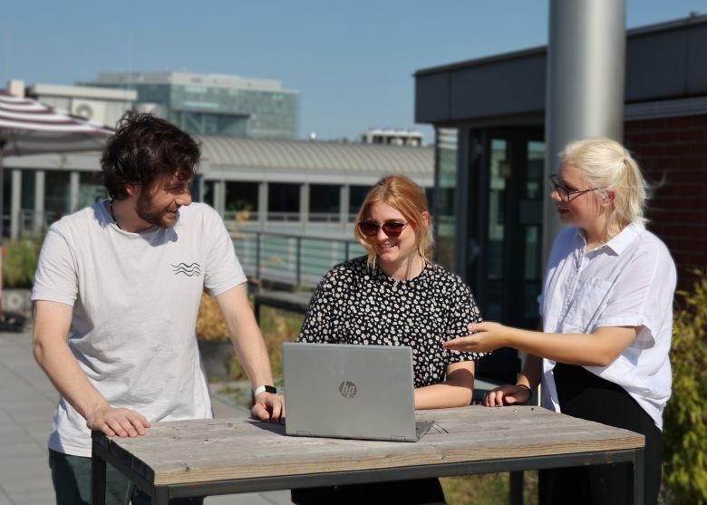 Ein Foto von einem jungen Mann und zwei jungen Frauen in T-Shirt auf einer Dachterasse, die gemeinsam an einem Tisch mit einem Laptop in der Sonne stehen