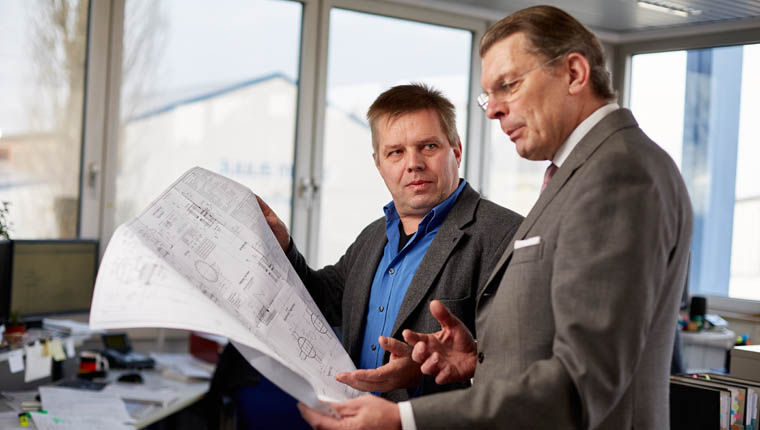 Zwei Manager stehen in einem Buero mit grossen Fenstern und schauen auf einen grossen Plan den beide in der Hand halten