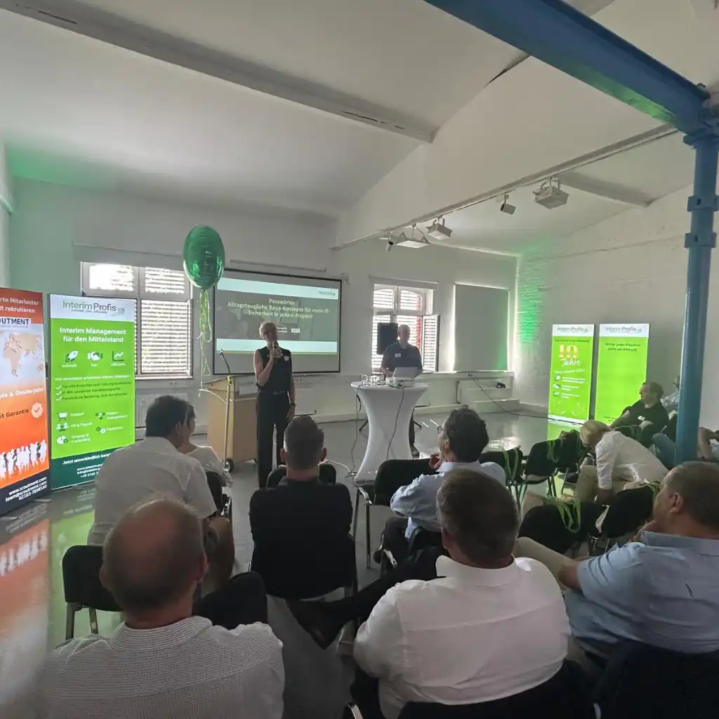 Aufnahme von unserem Interim Managerinnen und Managern die zu Gast des Jubiläums waren. Sitzend in einem großem Raum mit Annette Elias als Sprecherin neben ;agnus Barth. Daneben stehen Roll-Ups von den Interim Profis und SCOUTMENT
