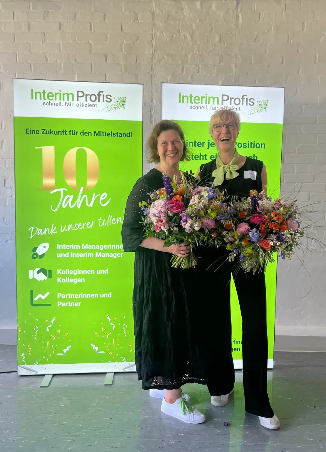 Zehn Jahre Interim Profis Kathrin Scholz-Arps und Annette Elias halten jeweils Blumensträße in der Hand und stehen vor zwei großen Roll-Ups